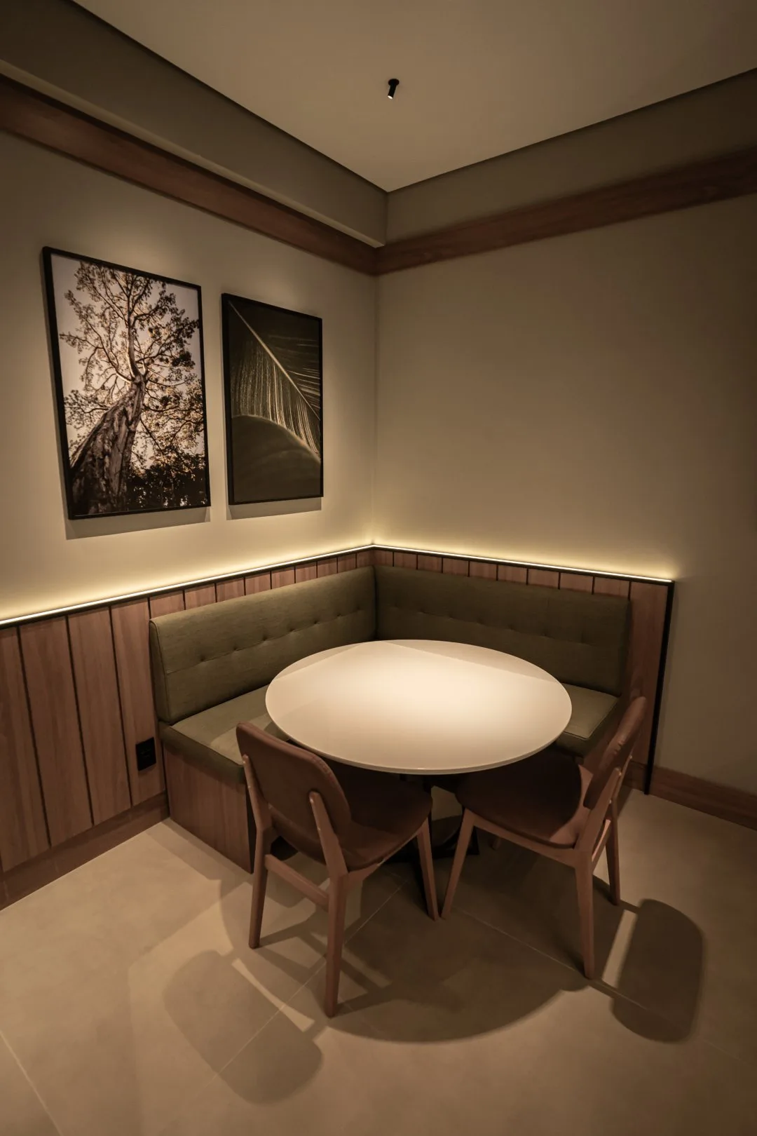 Canto com mesa redonda branca, banco estofado verde, cadeiras de madeira e dois quadros na parede