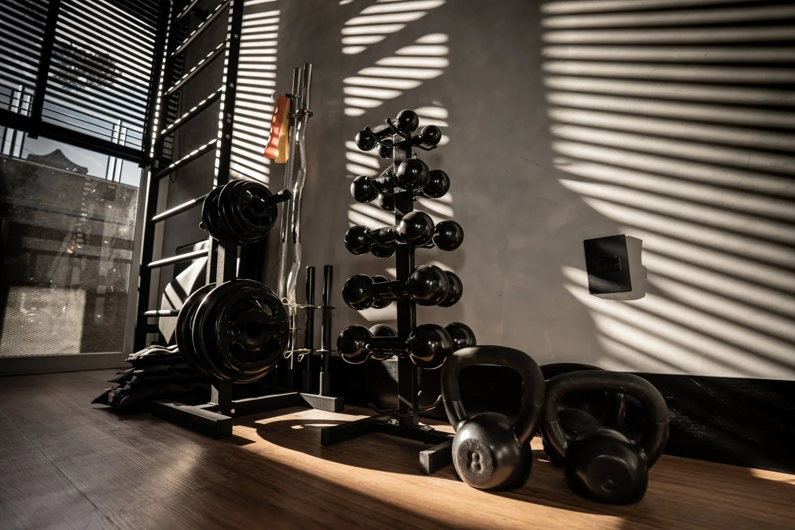 Espaço fitness com anilhas, suporte de halteres e kettlebells sob a luz filtrada pela persiana