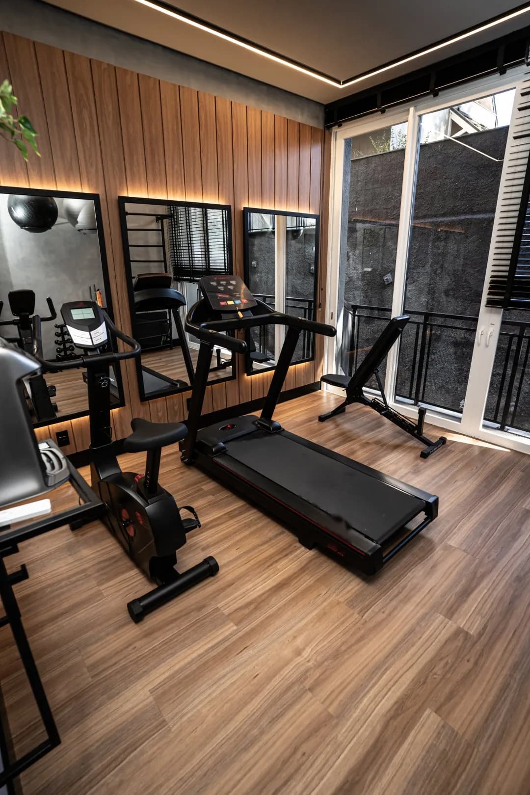 Espaço fitness com esteira, bicicleta ergométrica, banco de musculação, espelhos e porta de vidro para a sacada