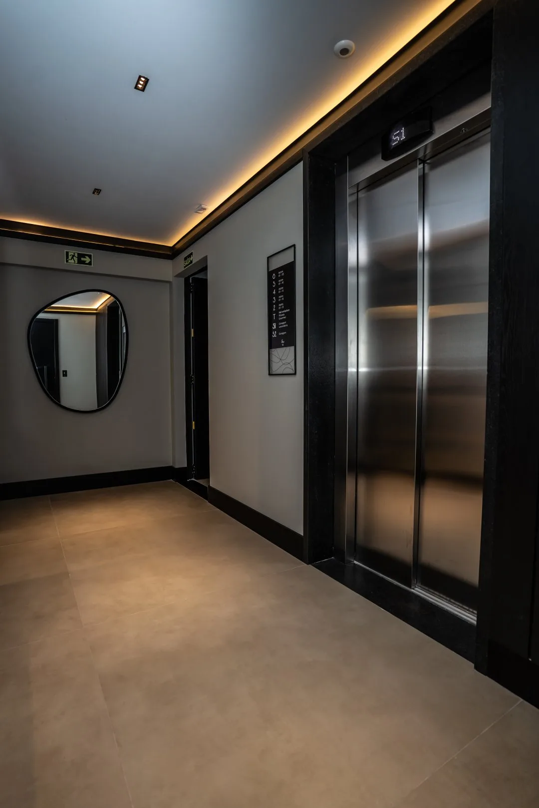 Hall do elevador com porta de inox, espelho de formato orgânico e iluminação indireta no forro