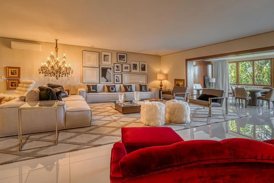 Sala de estar com sofás claros, lustre de cristal, parede de quadros e sofá de veludo vermelho em primeiro plano, com janelas para as árvores ao fundo