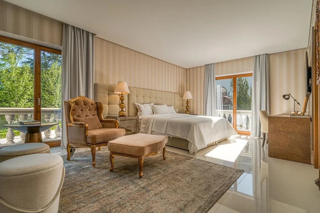 Suíte com papel de parede listrado, poltrona e pufe de estilo clássico, cama de casal e duas portas-janelas com vista para as árvores