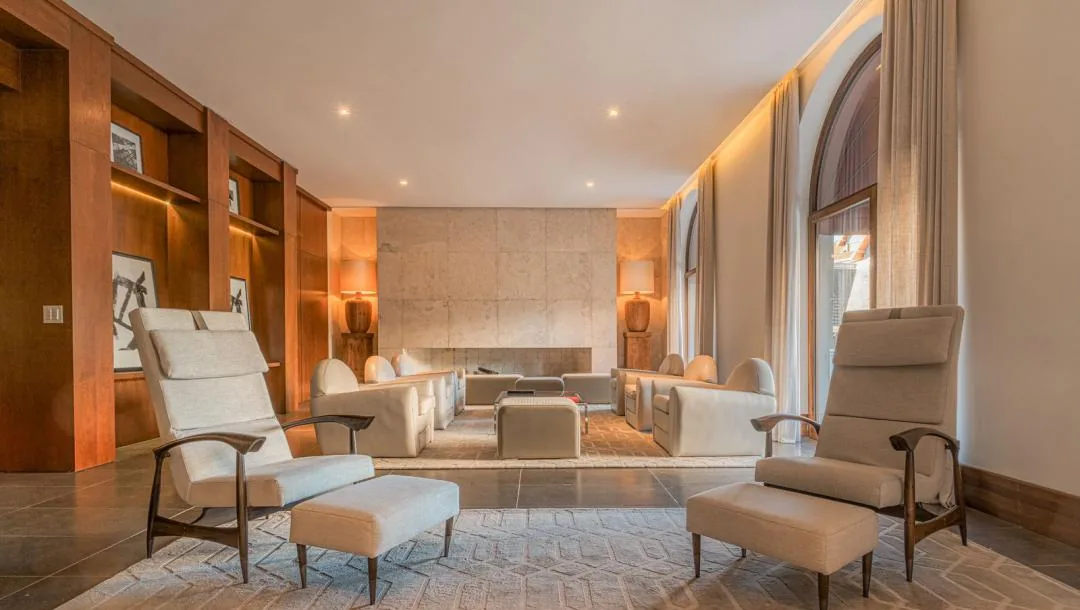Lounge com poltronas claras, estantes de madeira embutidas, parede de pedra ao fundo e janelas altas em arco