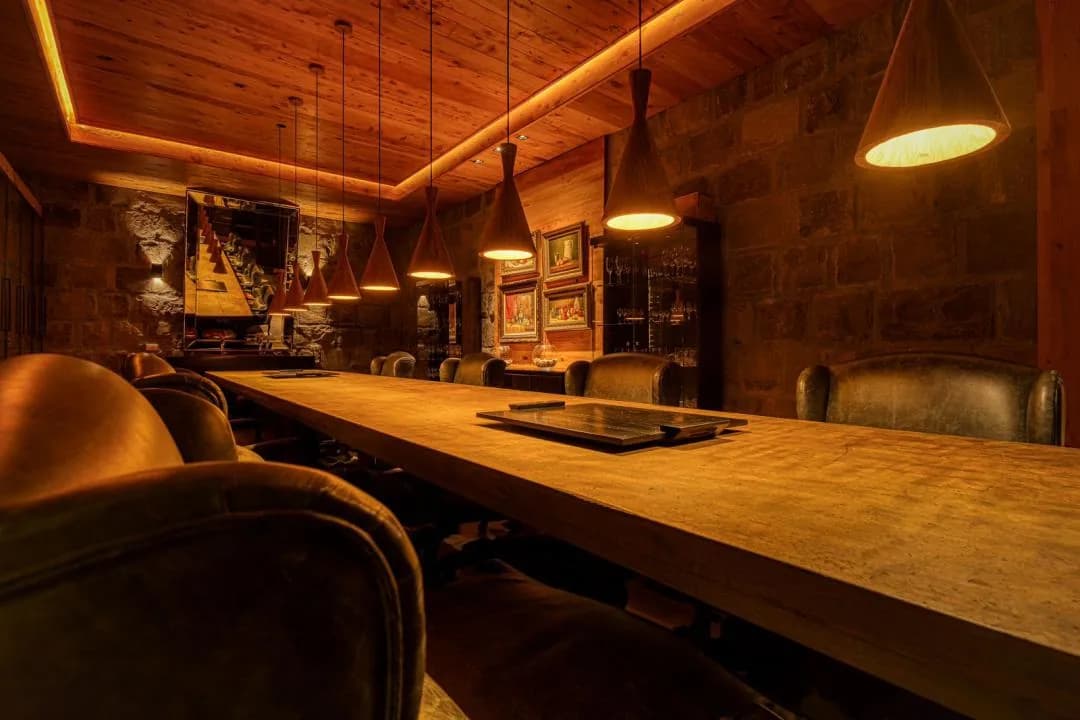 Sala de degustação com mesa longa de madeira, poltronas de couro, paredes de pedra, luminárias pendentes e armário com taças