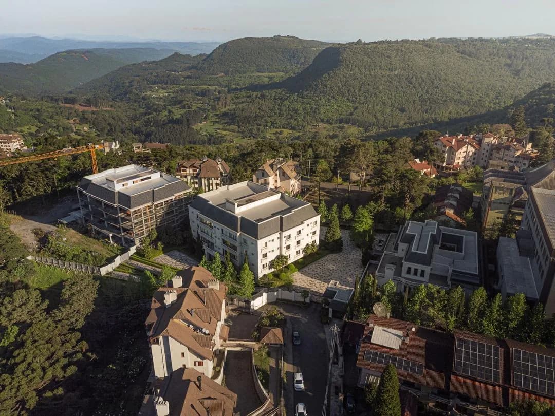 Vista aérea do bairro, com prédio branco de telhado cinza entre árvores e o vale com morros ao fundo