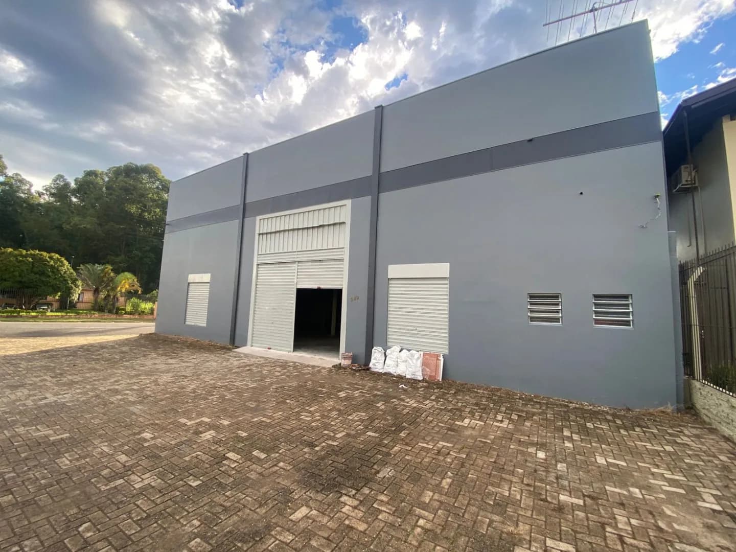 Fachada frontal com portão de enrolar aberto, duas aberturas com porta de aço, venezianas de ventilação e pátio de piso intertravado