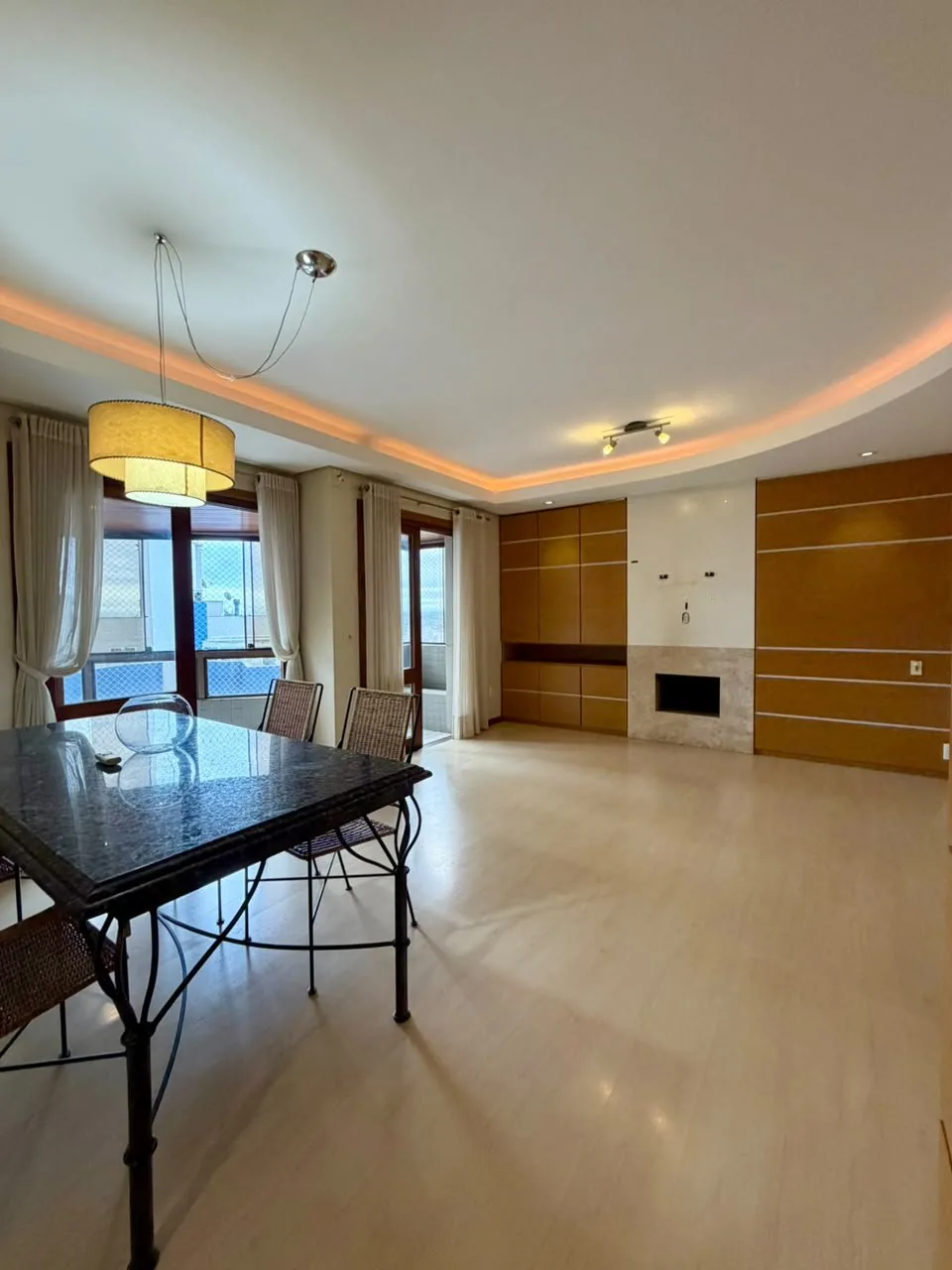 Sala com mesa de granito escuro, lareira de pedra clara entre painéis de madeira e janelas com cortinas
