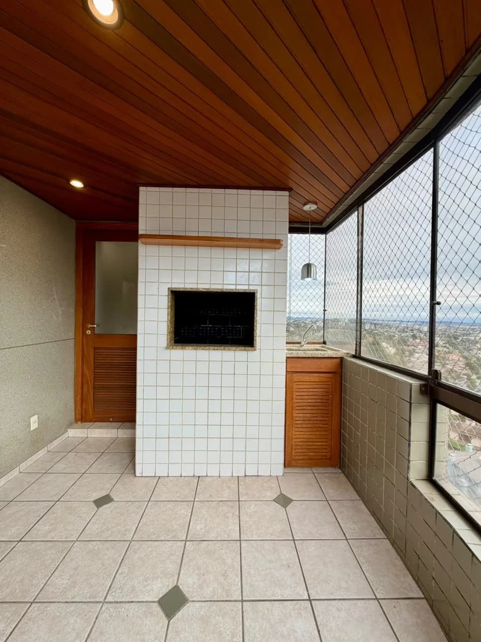 Sacada envidraçada com churrasqueira revestida de azulejo branco, bancada com armário de madeira e forro de madeira