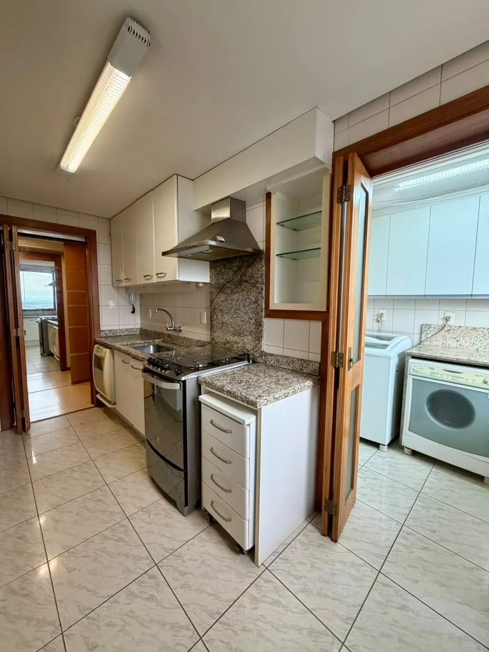 Cozinha com armários brancos, bancada de granito, fogão de inox com coifa e porta sanfonada aberta para a área de serviço
