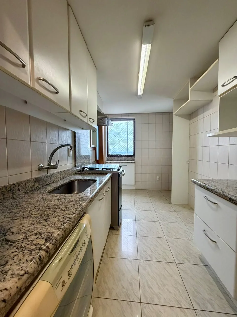 Cozinha em corredor com armários brancos, bancada de granito com cuba de inox e janela ao fundo