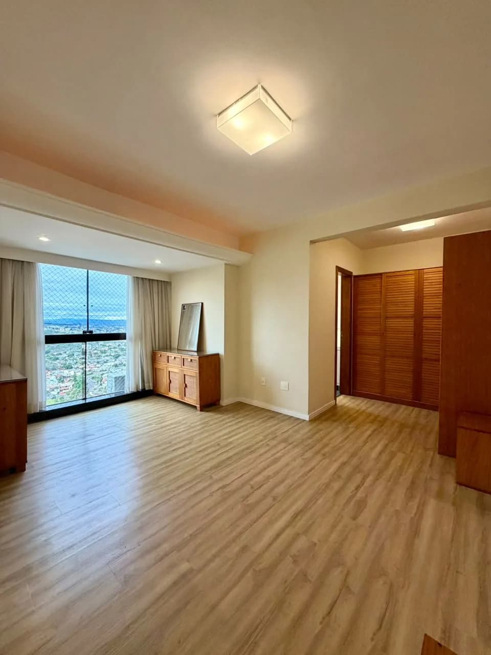 Ambiente com piso vinílico amadeirado, janela ampla com vista da cidade, aparador de madeira e armário com portas venezianas