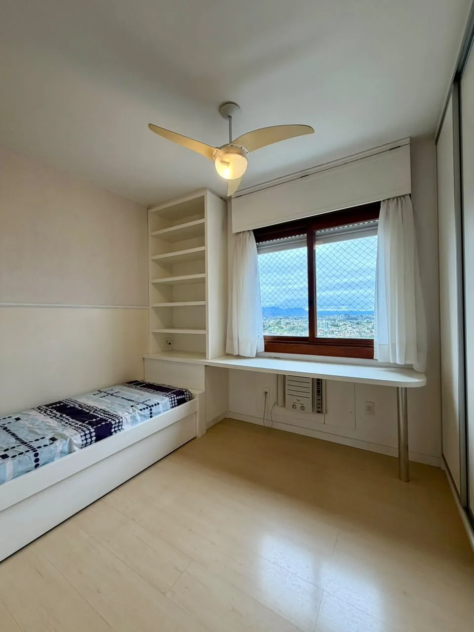 Quarto com cama de solteiro, estante e escrivaninha embutidas, ventilador de teto e janela com vista