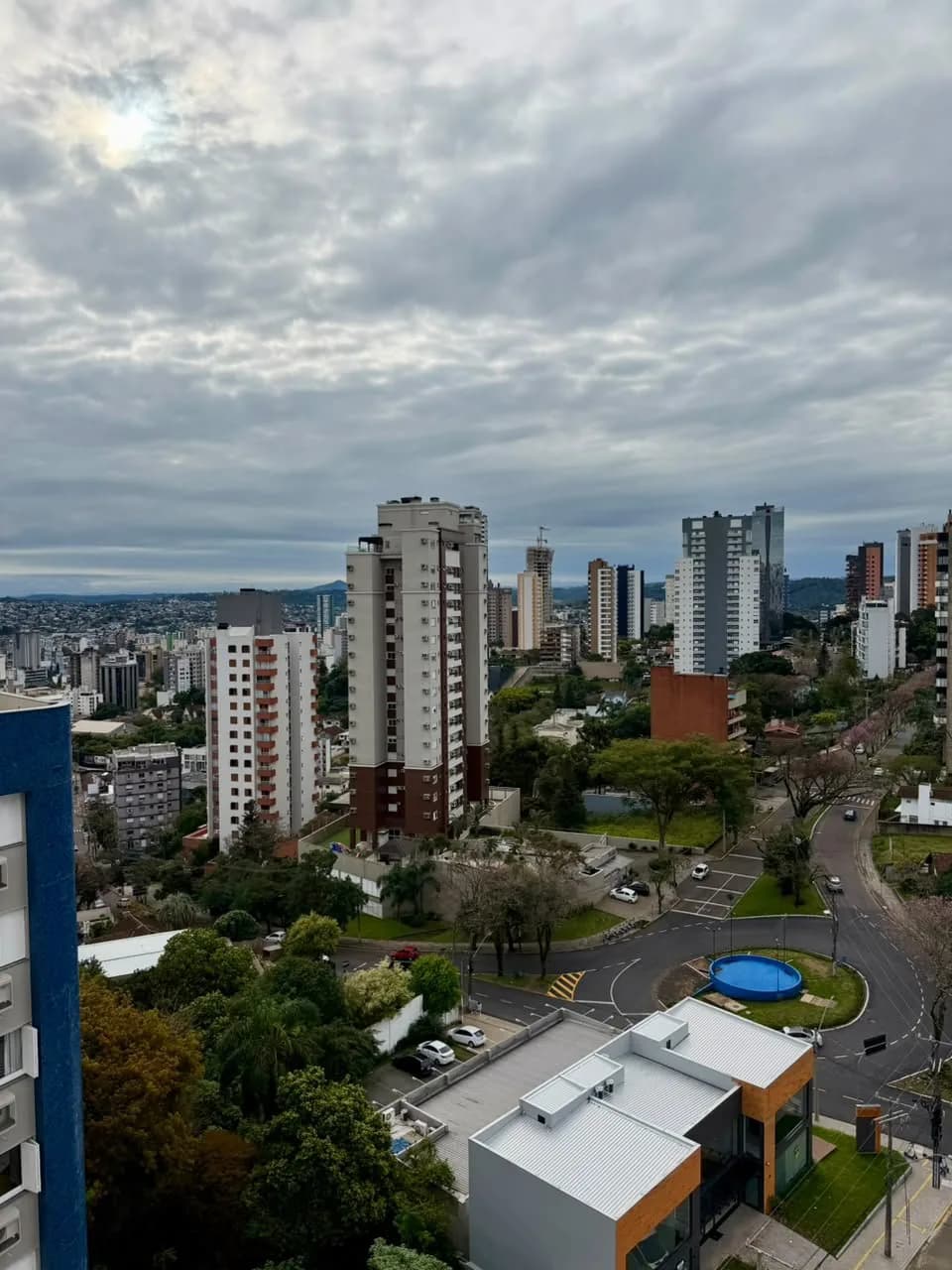 Vista do alto para prédios residenciais, rotatória arborizada e morros ao fundo