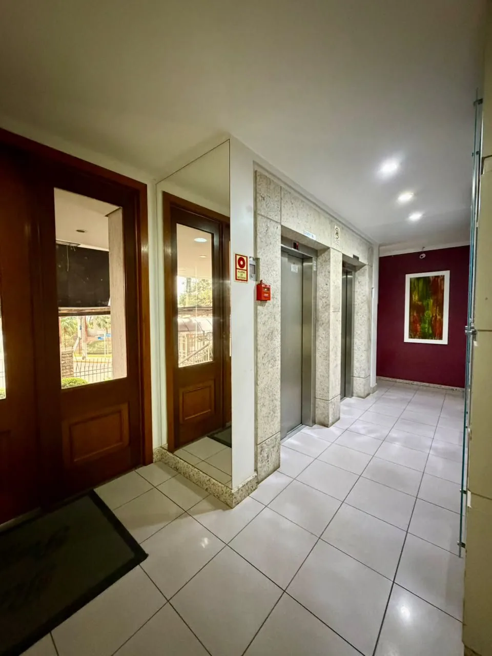 Hall de entrada do prédio com dois elevadores, piso cerâmico claro e portas de madeira com vidro