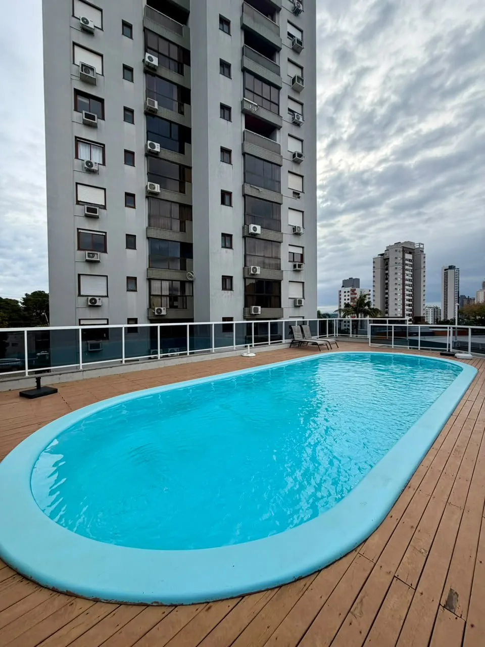 Piscina do condomínio sobre deck de madeira, com guarda-corpo de vidro e espreguiçadeiras ao fundo