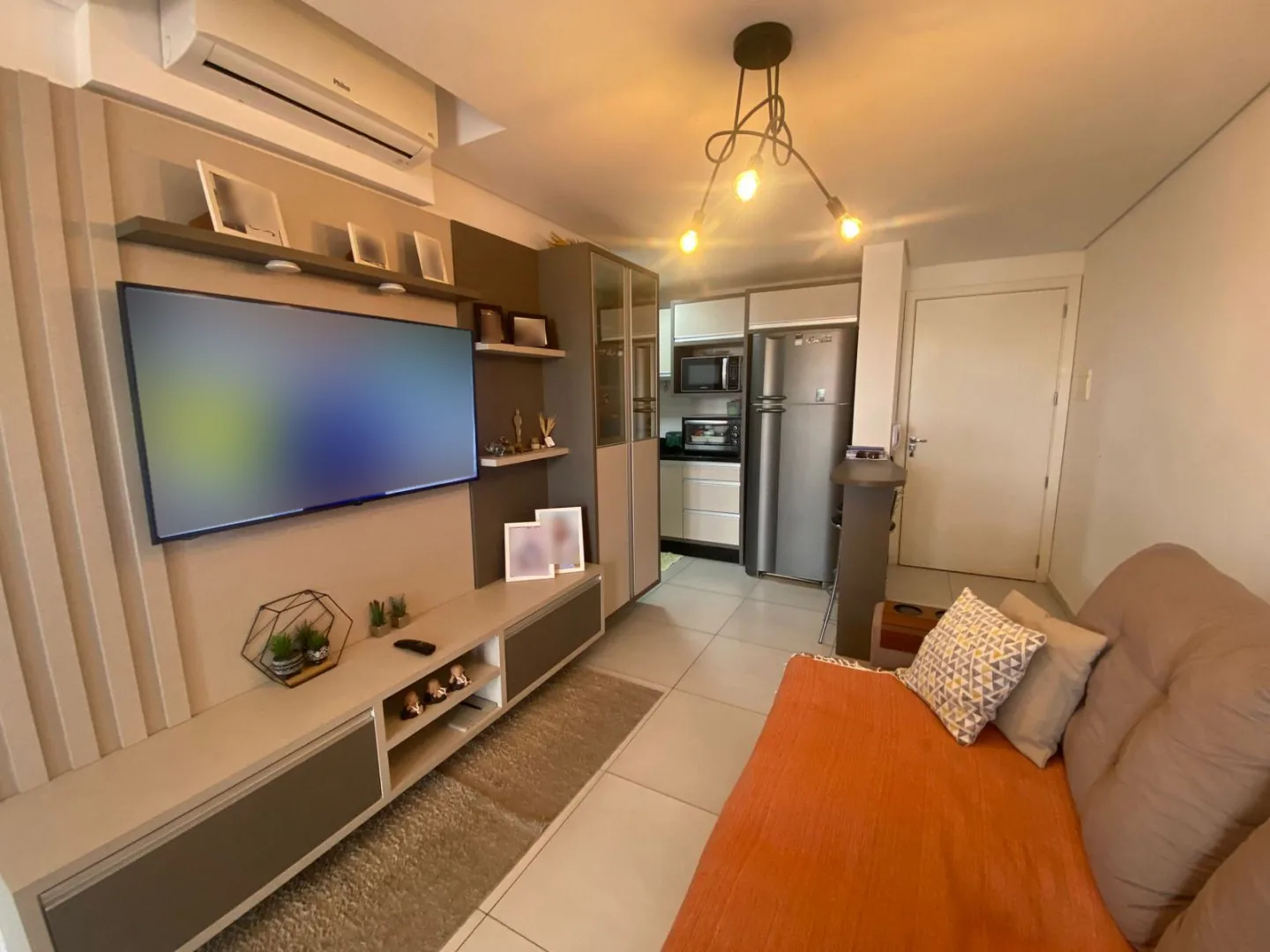 Sala de estar com painel de TV em tons de bege, sofá com manta laranja, luminária pendente de lâmpadas aparentes e cozinha integrada ao fundo