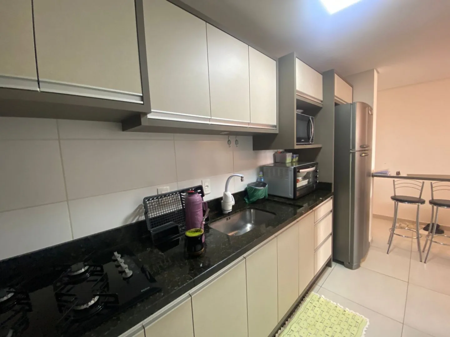 Cozinha com cooktop, pia de inox em bancada de granito preto, geladeira inox e balcão com banquetas ao fundo