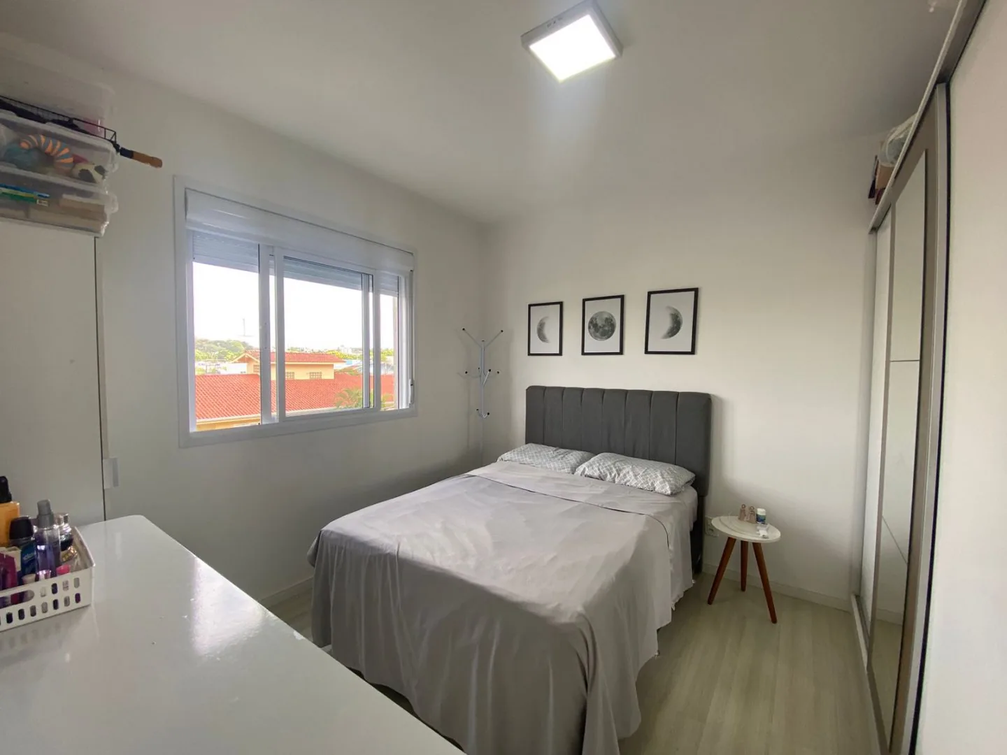 Dormitório com cama de casal, cabeceira estofada cinza, janela ampla e roupeiro com porta de espelho