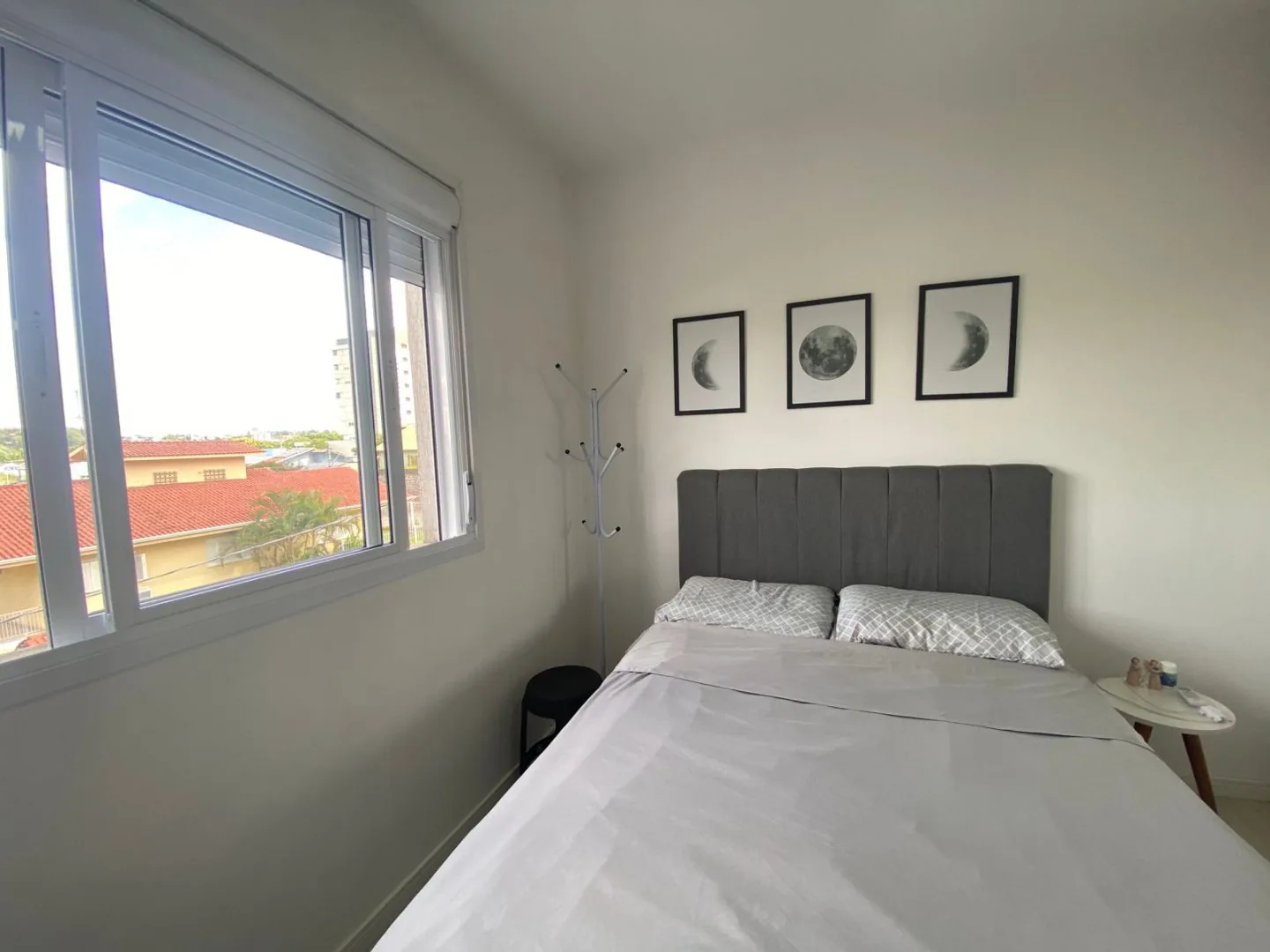 O mesmo dormitório visto de perto, com três quadros de lua sobre a cama e janela com vista para os telhados vizinhos