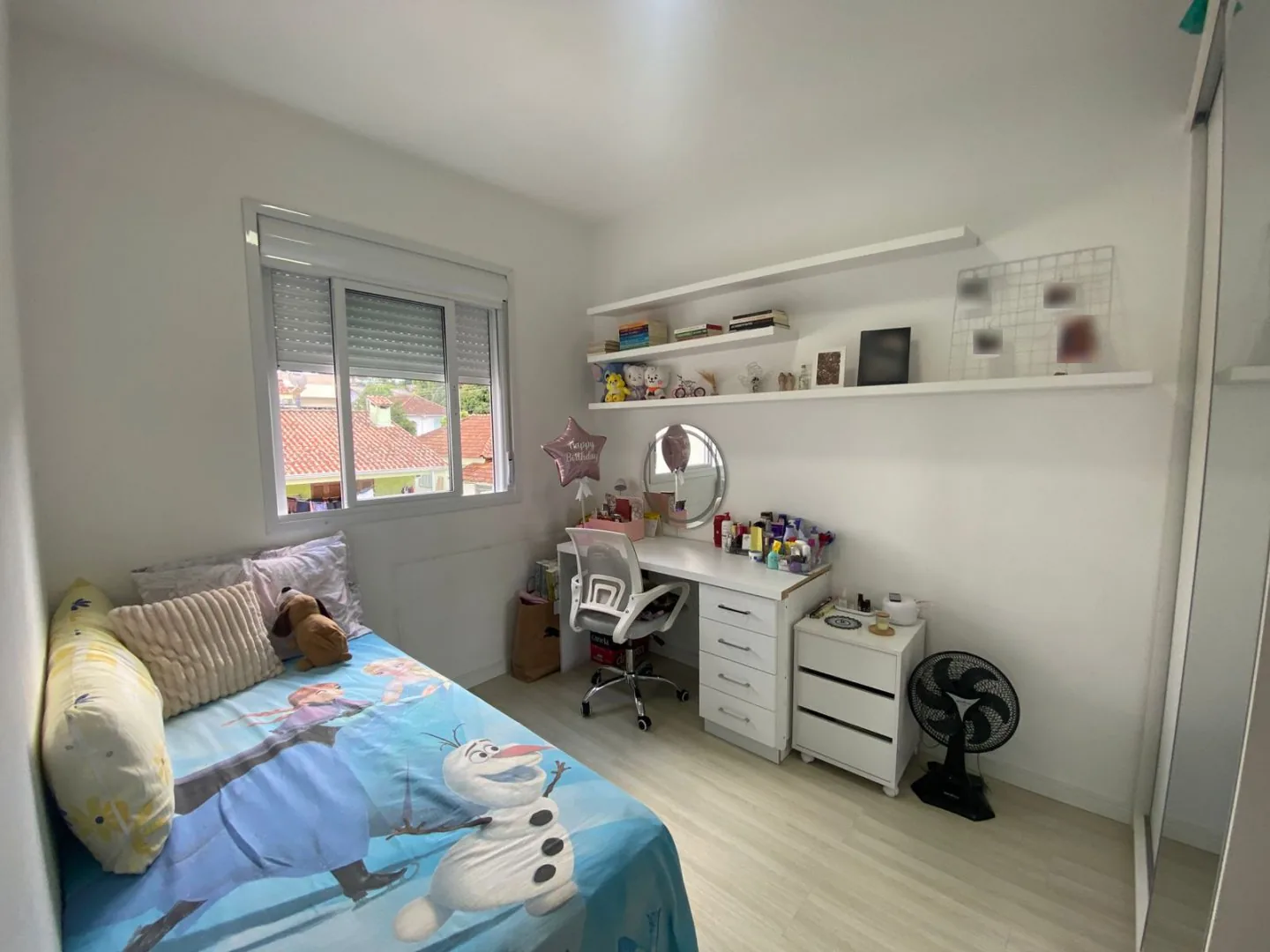 Dormitório com cama, escrivaninha branca com gaveteiro, prateleiras suspensas e janela com persiana
