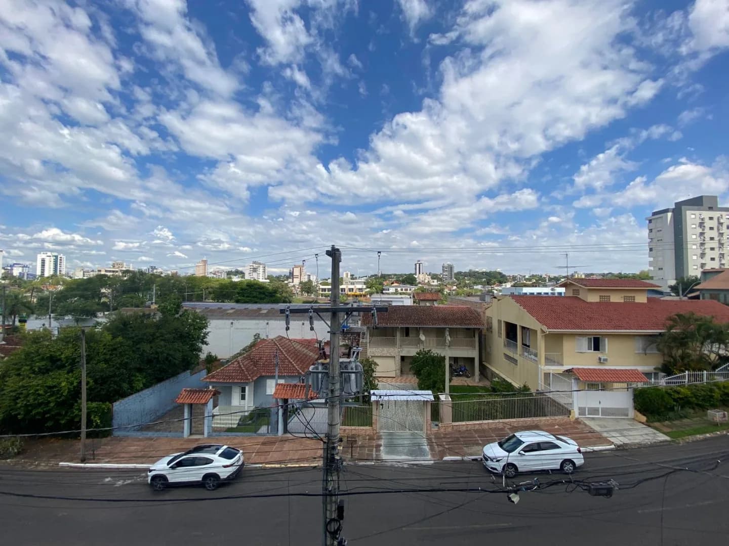 Vista da sacada para a rua e as casas do bairro, com prédios da cidade ao fundo sob céu azul