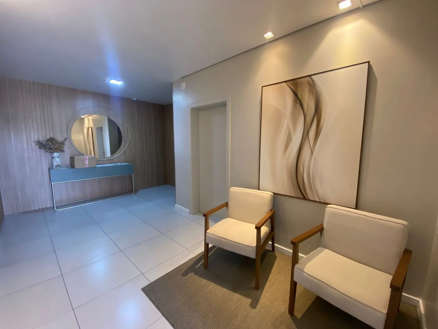 Hall do prédio com duas poltronas claras, quadro abstrato grande e piso de porcelanato