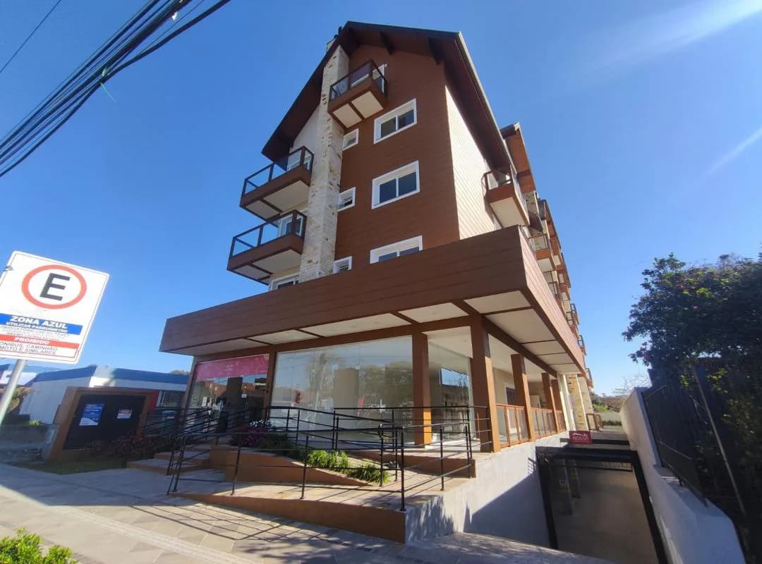 Fachada do prédio com revestimento amadeirado, coluna de pedra, sacadas com guarda-corpo de vidro, telhado inclinado e lojas envidraçadas no térreo