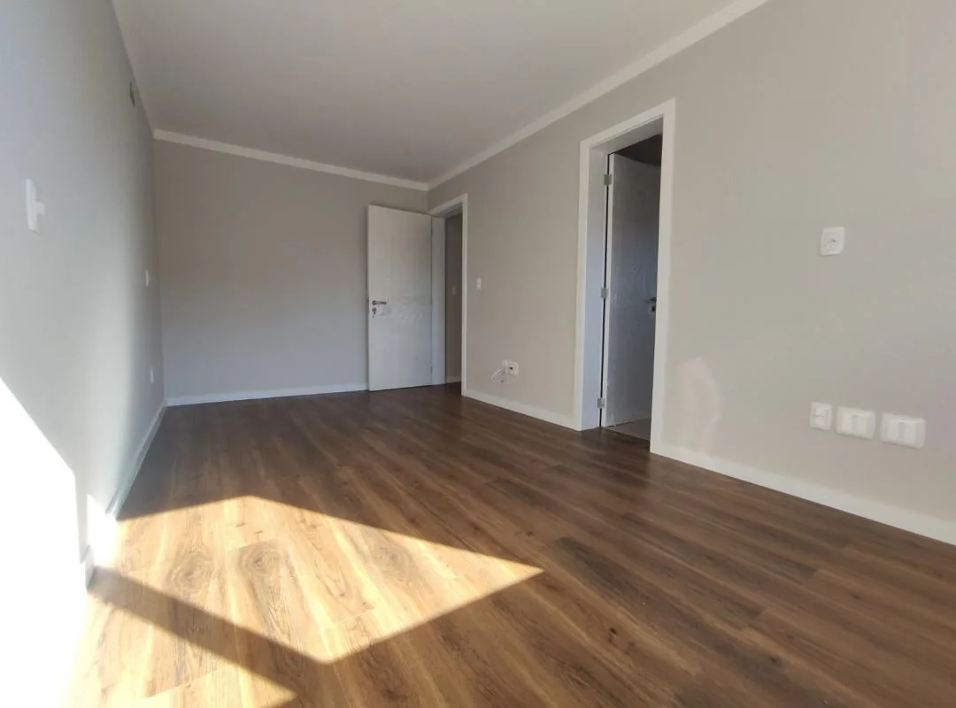 Quarto com piso vinílico amadeirado, paredes cinza-claro, duas portas brancas e sol batendo no piso