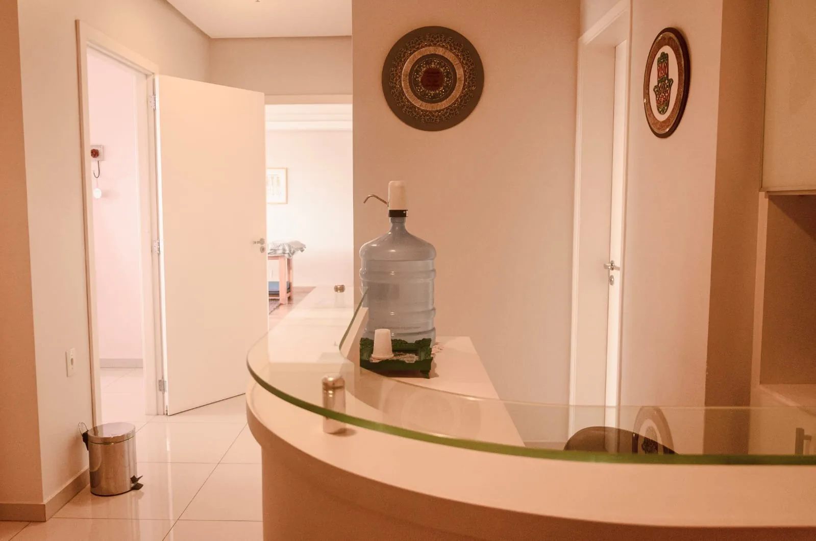Recepção com balcão curvo de tampo de vidro, porta aberta e passagem para a sala com maca ao fundo