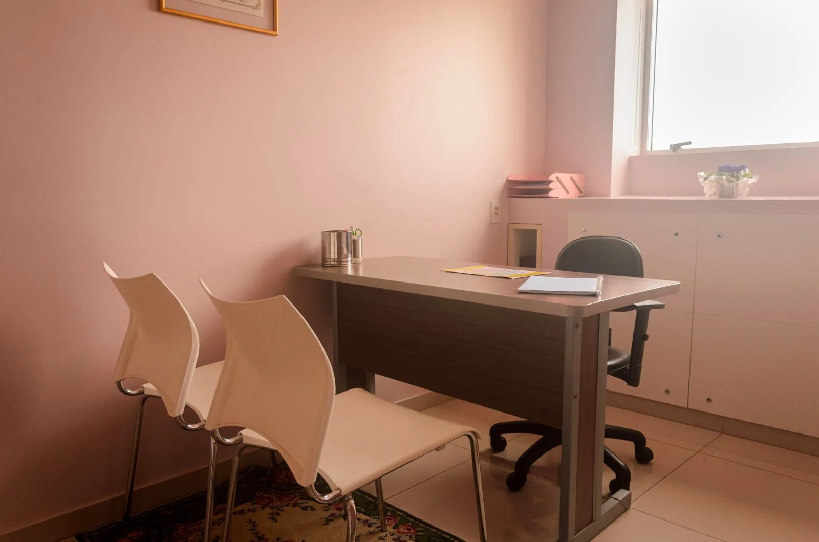 Sala com mesa de escritório, duas cadeiras brancas e armário baixo sob a janela