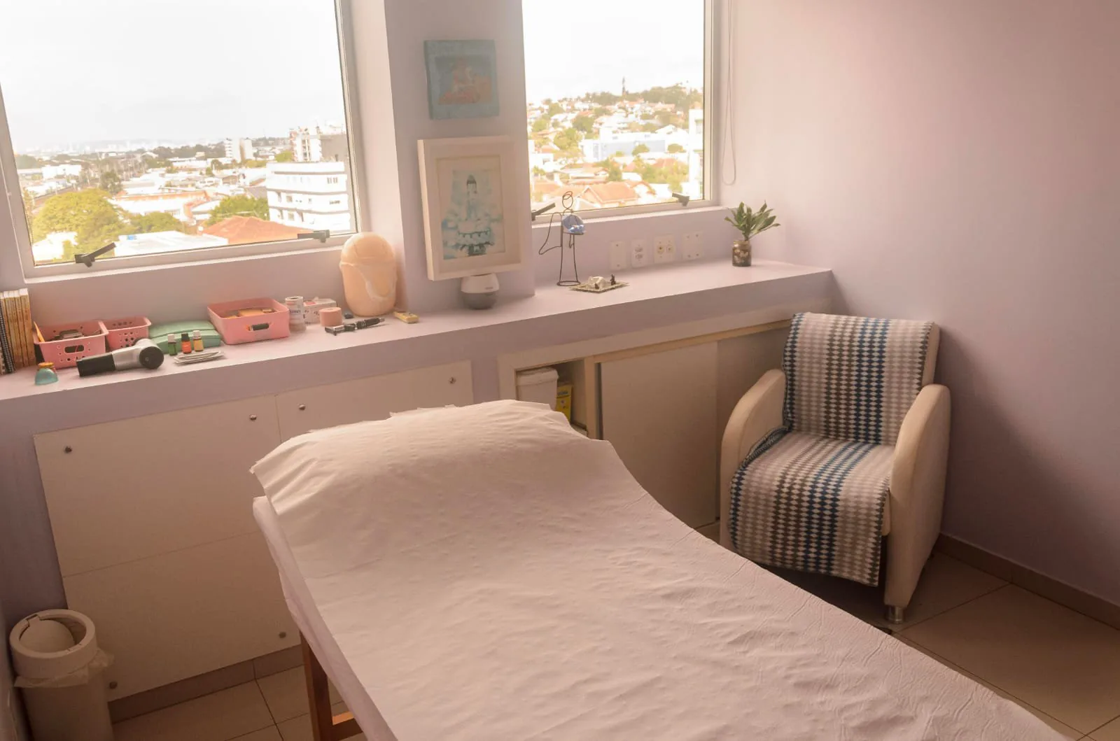 Sala com maca, poltrona, bancada branca sob a janela e vista para os telhados e prédios do bairro