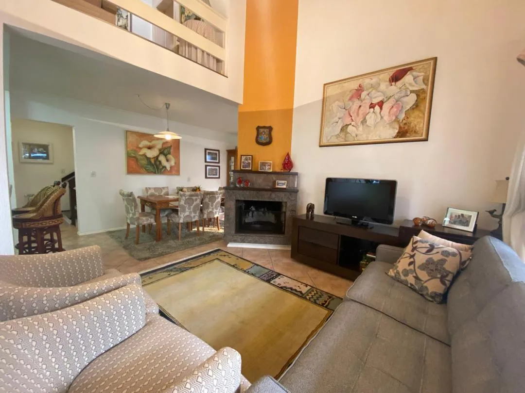 Sala de estar com pé-direito alto, lareira, sofá cinza, poltronas estampadas e mesa de jantar ao fundo