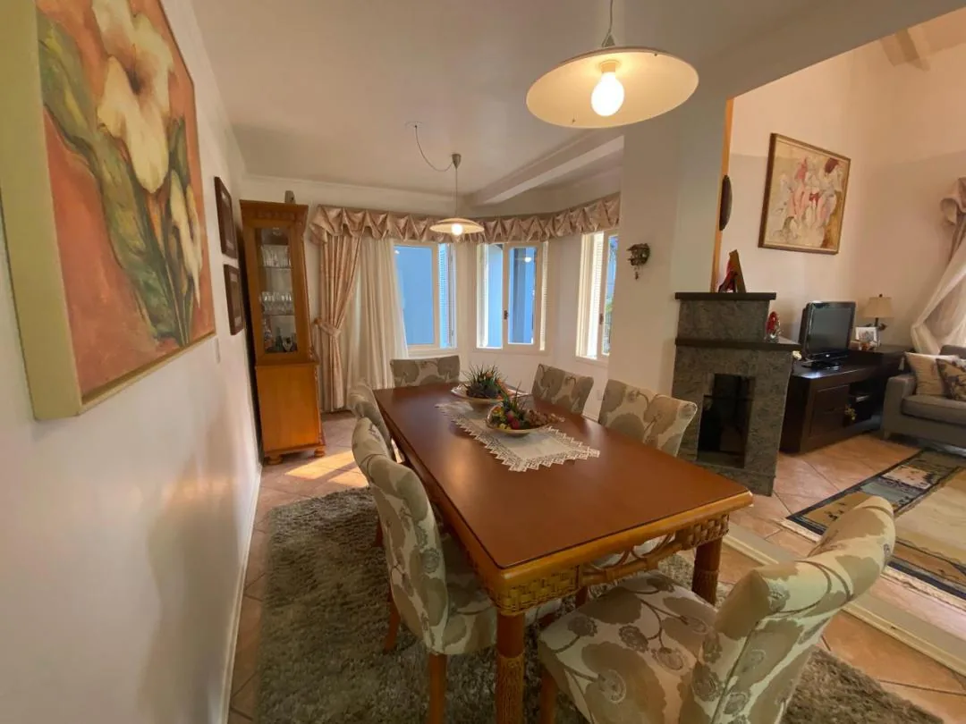 Sala de jantar com mesa de madeira, cadeiras estofadas estampadas, cristaleira e três janelas em ângulo ao fundo