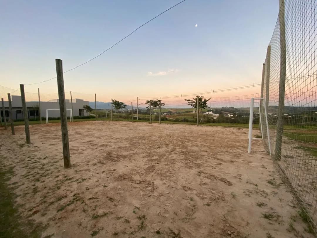 Quadra de areia cercada por rede alta, com traves de futebol e morros ao fundo