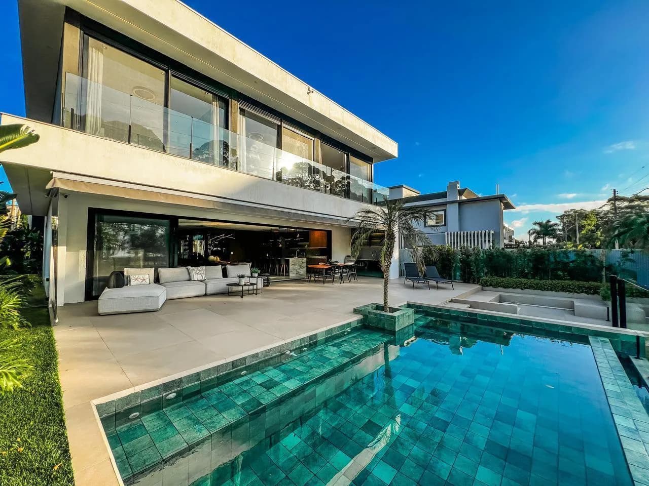 Fundos da casa de dois pavimentos com piscina de revestimento verde, palmeira em ilha e varanda integrada à sala