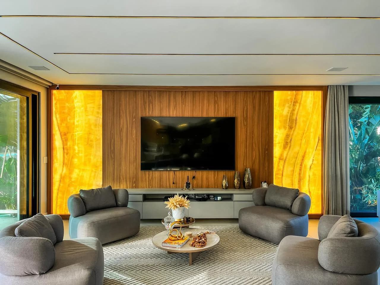 Sala de TV com painel de madeira entre dois painéis de ônix iluminados e quatro poltronas cinza