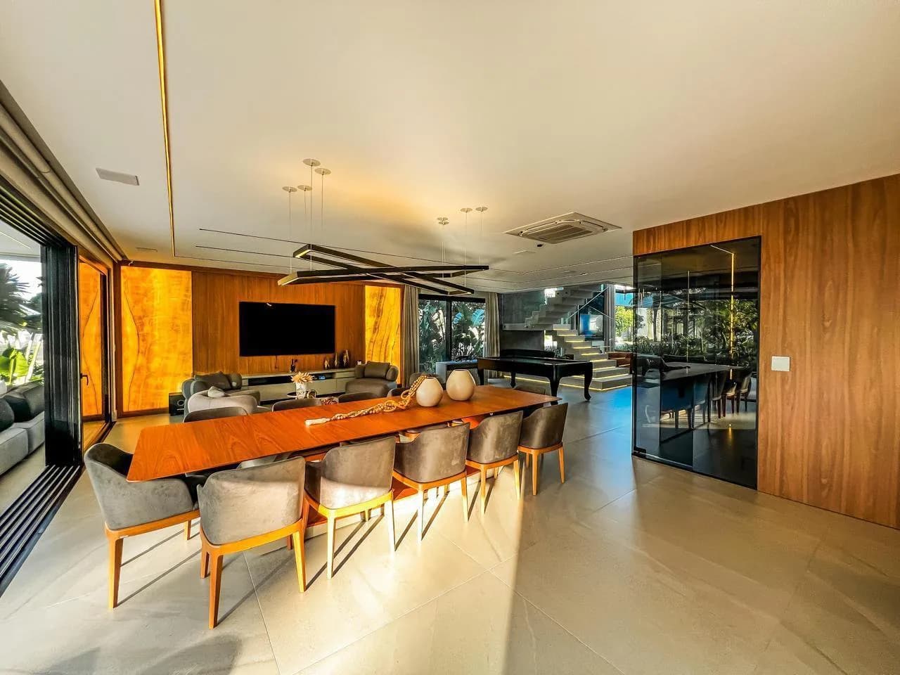Mesa de jantar longa de madeira com cadeiras estofadas, mesa de sinuca e escada iluminada ao fundo
