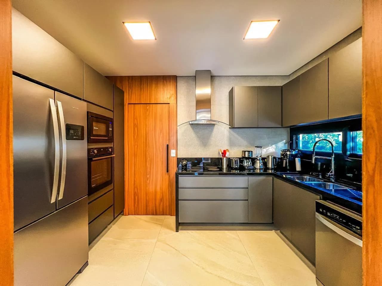 Segunda cozinha com armários cinza, bancada de granito preto, geladeira inox de duas portas e forno embutido