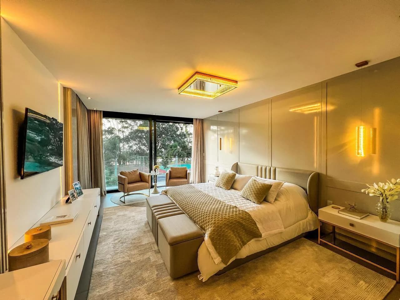 Suíte com cama de casal, calçadeira, par de poltronas junto à janela de piso a teto e TV na parede
