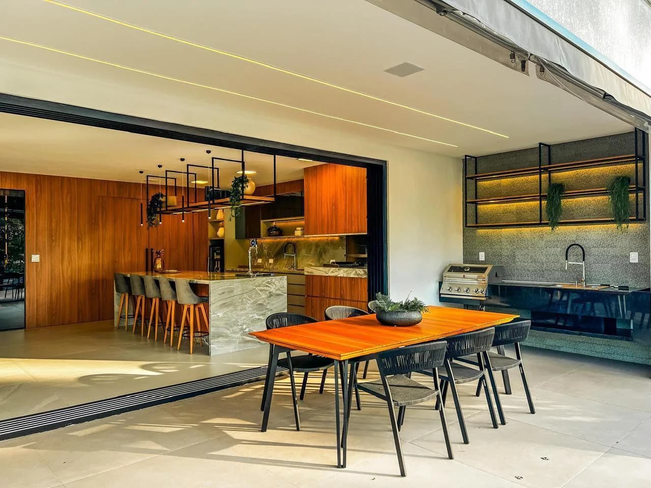 Área gourmet externa com mesa de madeira e seis cadeiras, churrasqueira de inox e bancada com pia, aberta para a cozinha