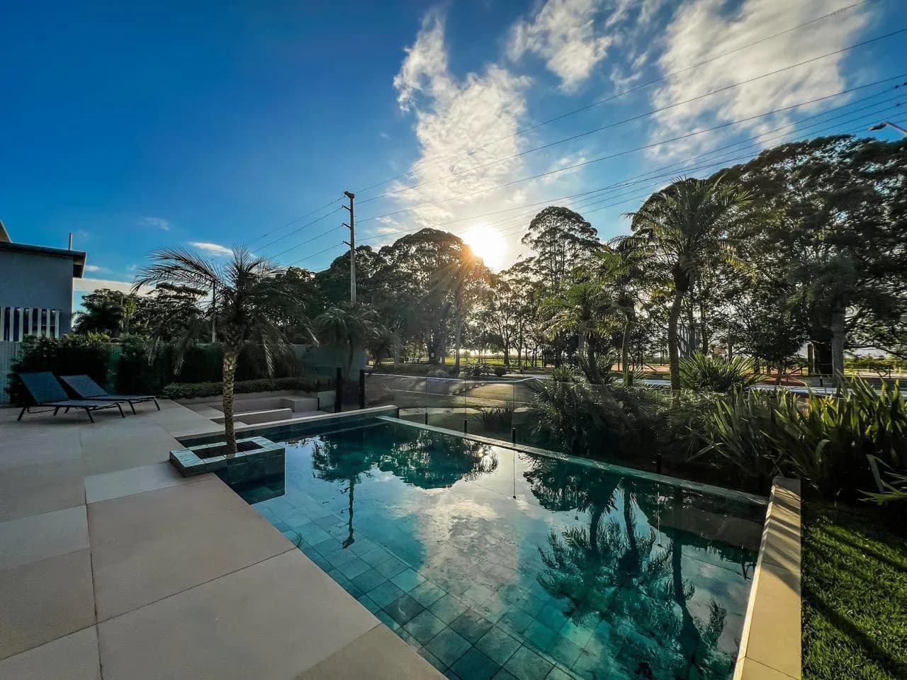 Piscina com sol baixo, espreguiçadeiras, palmeira e árvores altas ao fundo
