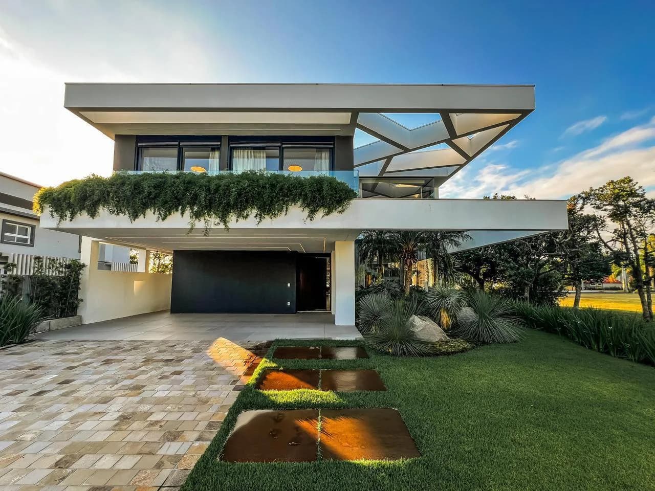 Fachada frontal branca com pérgola triangular vazada, floreira pendente na sacada, garagem e caminho de pisantes no gramado