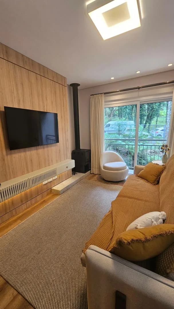 Sala com painel de madeira, TV na parede, lareira a lenha preta, poltrona bege e porta de vidro com árvores do lado de fora