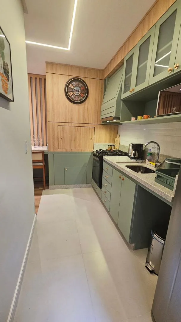 Cozinha em corredor com armários verdes, painel de madeira com relógio de parede e piso claro