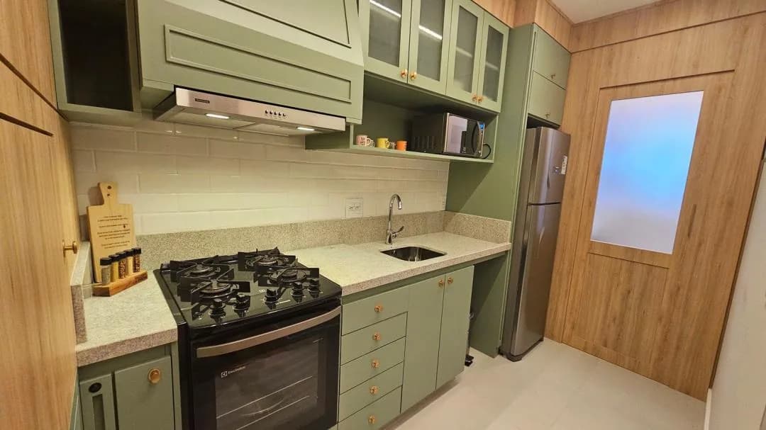 Cozinha com armários verdes, fogão, pia de inox em granito, geladeira e porta de madeira com vidro fosco