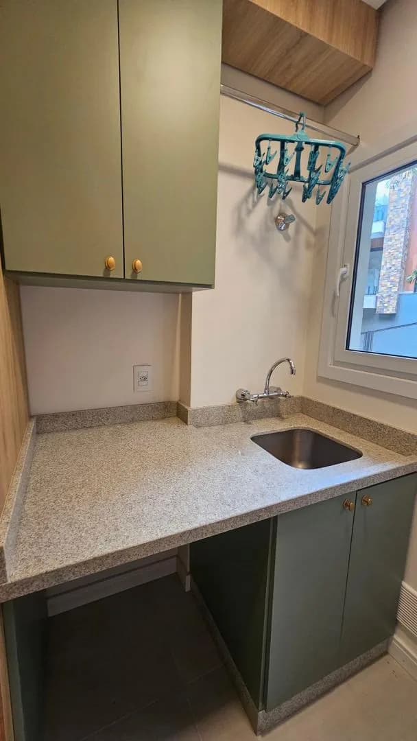 Área de serviço com cuba de inox em bancada de granito, armário verde e janela de PVC