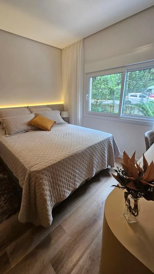 Quarto com cama de casal, cabeceira iluminada e janela de correr com vista para árvores