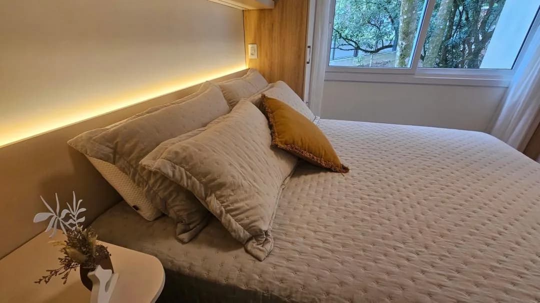 Cama de casal com cabeceira em painel, fita de luz quente e almofadas, com janela ao lado