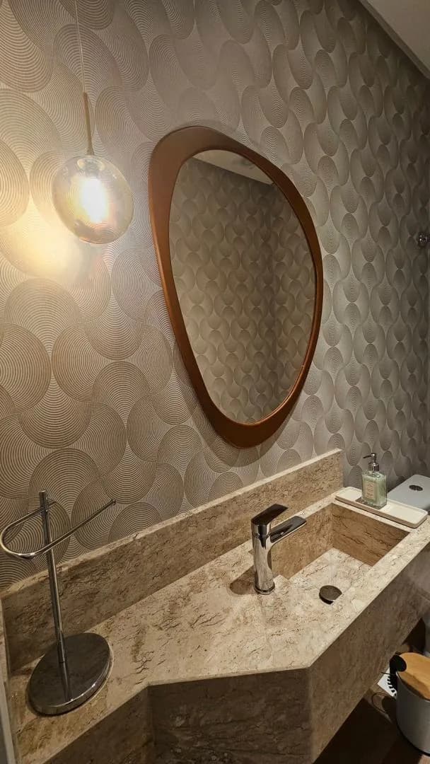 Lavabo com cuba esculpida em pedra, espelho de moldura de madeira em formato orgânico e papel de parede texturizado