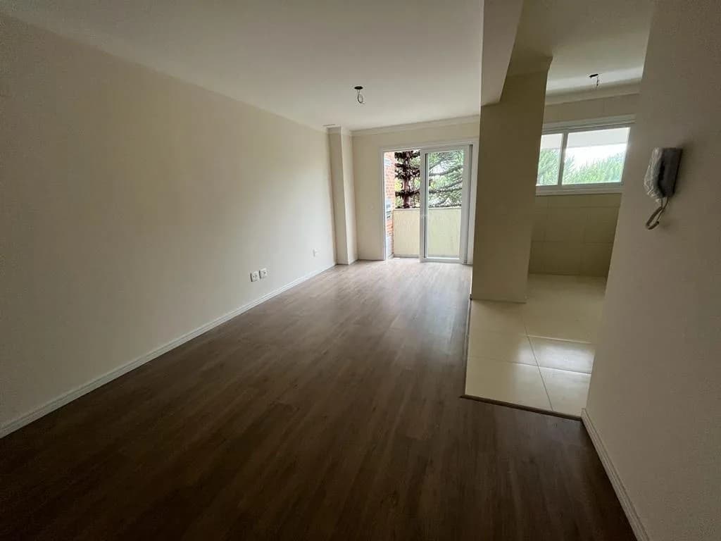 Sala ampla e vazia com piso laminado escuro, cozinha integrada à direita e porta de vidro para a sacada ao fundo
